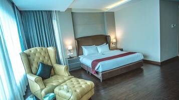 A hotel room with a large bed, a tufted armchair, a glass side table, and a nightstand with lamps.