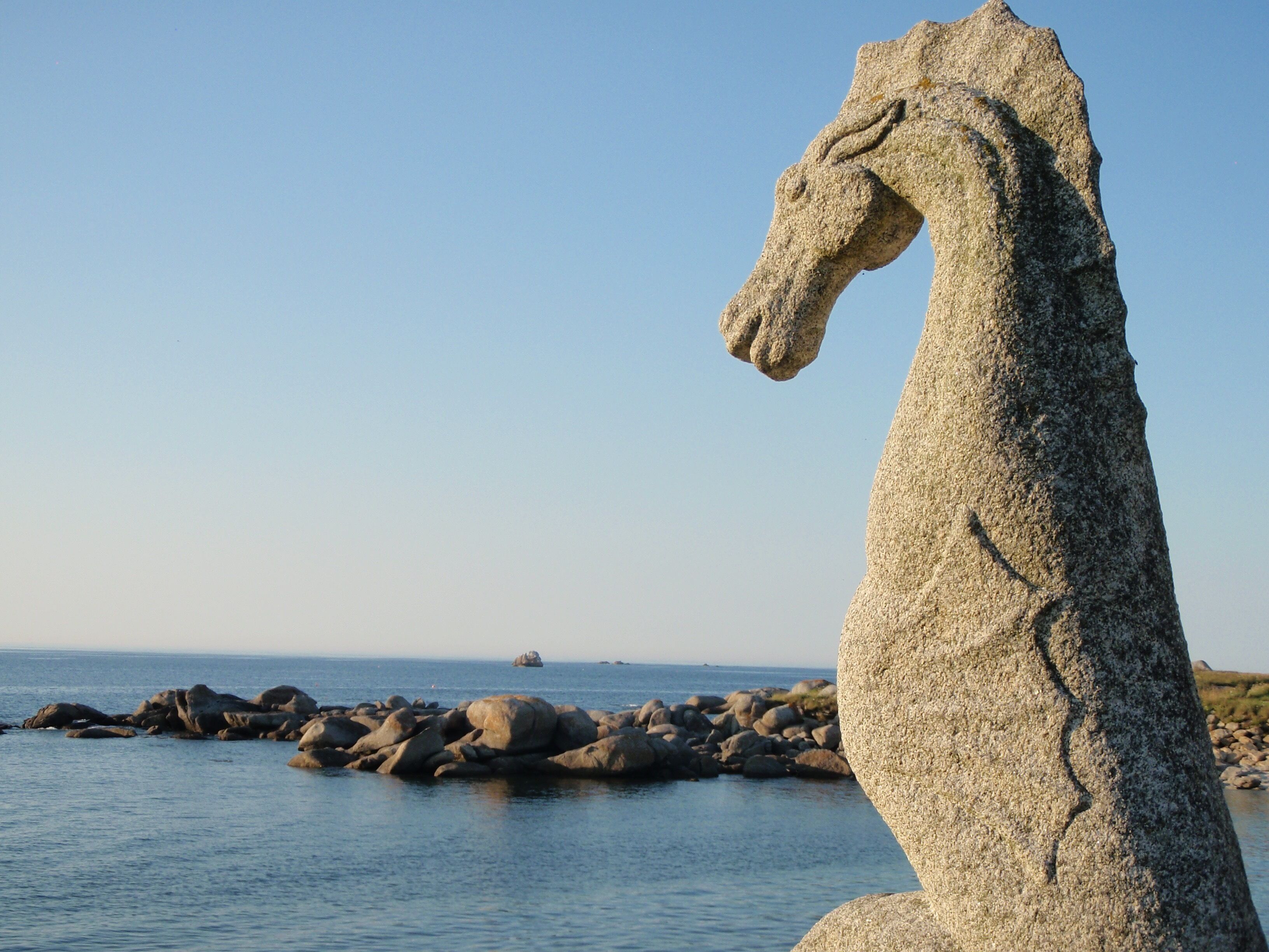 A stone horse statue by the sea with a rocky shoreline and calm waters.