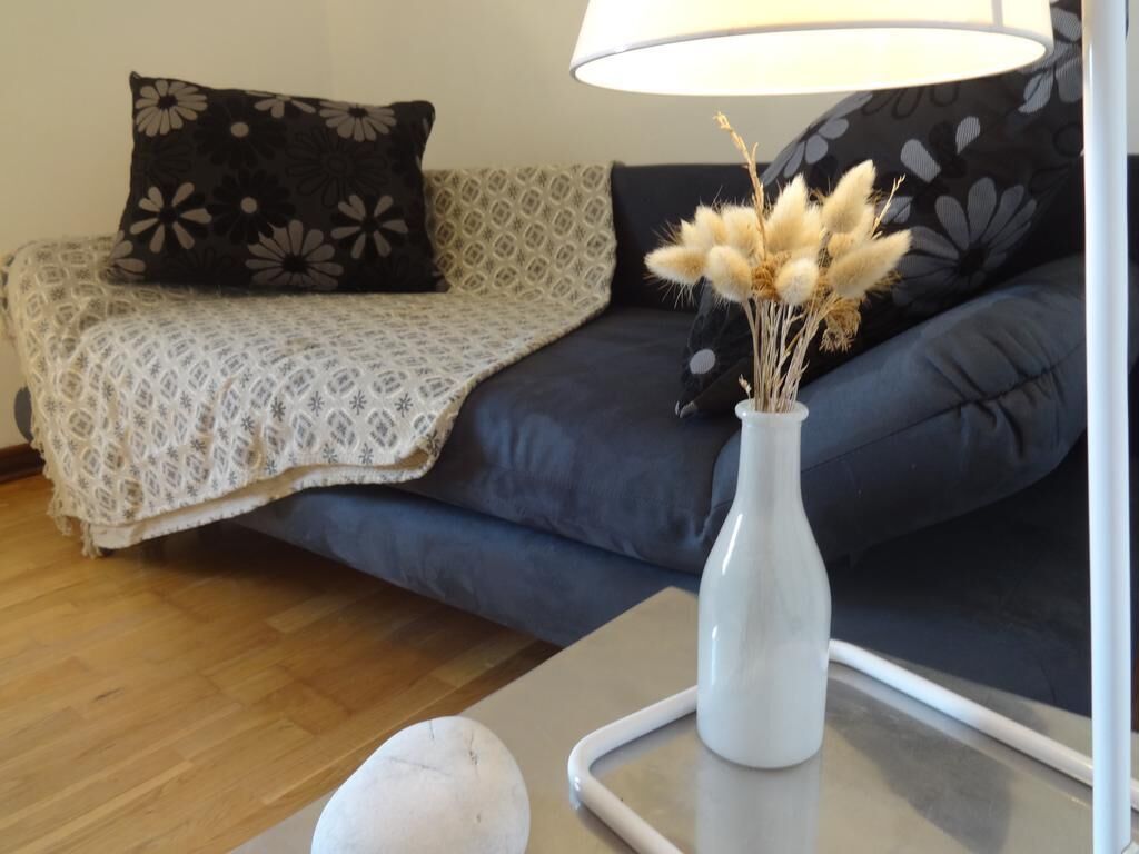 Een woonkamer met een bank, een geborduurd plaid, een witte vaas met gedroogde bloemen en een lamp.
