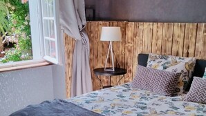 Een slaapkamer met een groot bed, een nachtkastje met een lamp, een rieten hanglamp en uitzicht op groen buiten.
