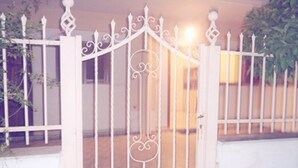Une porte en métal blanc, ornée de motifs décoratifs, qui donne accès à un bâtiment dont le mur est de couleur beige et qui possède une fenêtre.