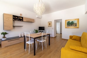 A modern living room with a wooden dining table, white chairs, a yellow sofa, a TV, and a chandelier.