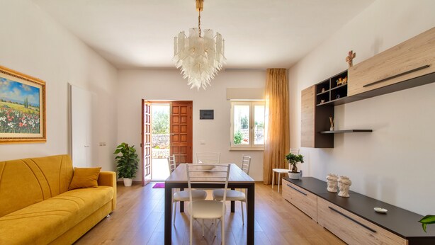 A modern living room with a yellow sofa, a dining table with chairs, a chandelier, and a kitchen area with wooden cabinets and a black countertop. - Pet-Friendly Apartment 'I due ulivi' with view on the hills of Modica, Terrace & Wi-Fi (Modica)