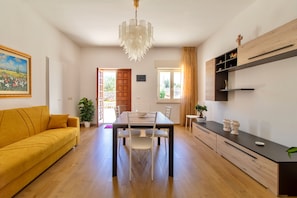 A modern living room with a yellow sofa, a dining table with chairs, a chandelier, and a kitchen area with wooden cabinets and a black countertop.