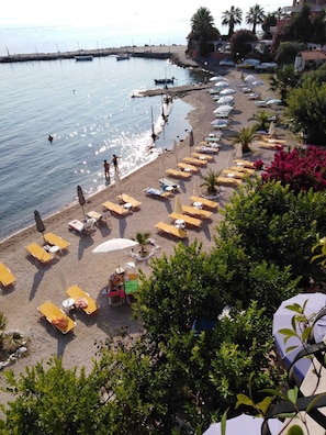 A beach with sun loungers and umbrellas, a pier with boats, and a building with a red roof.