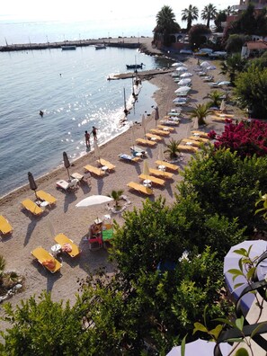 A beach with sun loungers and umbrellas, a pier with boats, and a building with a red roof.
