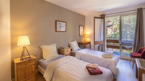 A hotel room with two beds, a nightstand with a lamp, a small table, and a chair. There is a window with curtains and a view of greenery outside.