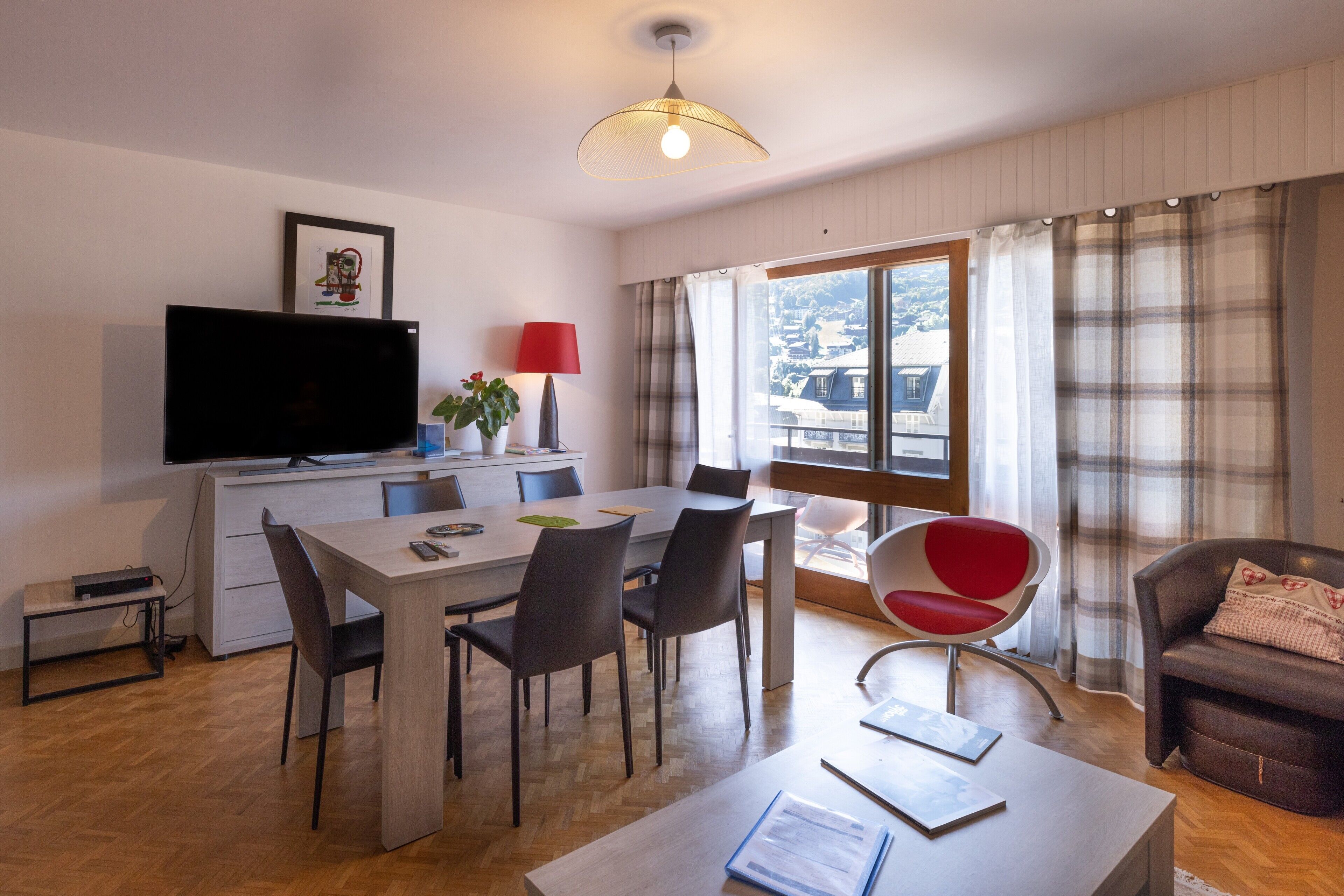 Una sala de estar moderna con comedor, televisor de pantalla plana, lámpara roja y vista a la ciudad a través de amplios ventanales.