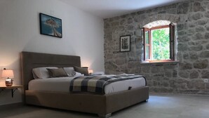 A bedroom with a stone wall, a bed with a headboard, a nightstand, a lamp, and a window with a view of greenery. - Private Stone House Next to the sea (Kotor)