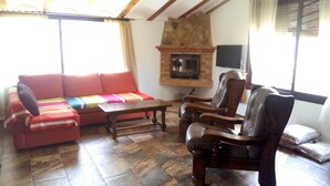 Ein Wohnzimmer mit einer Couch, zwei Sesseln, einem Holztisch, einem Steinkamin und einem an der Wand befestigten Fernseher.