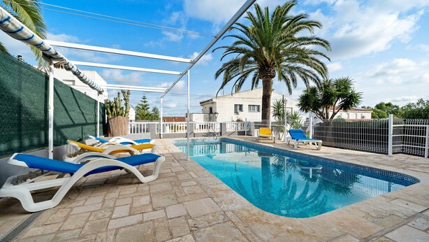 En pool med solstolar, en pergola, palmer och ett inhägnat område.