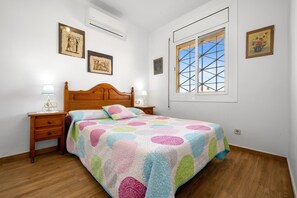 Ein Schlafzimmer mit einem hölzernen Bett, einem Nachttisch mit Lampe, einem Fenster mit Gittermuster und Wandbildern.