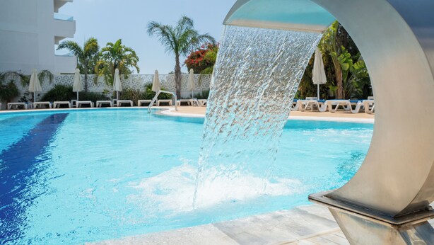 A swimming pool with a water feature, surrounded by lounge chairs and palm trees. - Gaia Sun N Blue Hotel (Ayia Napa)