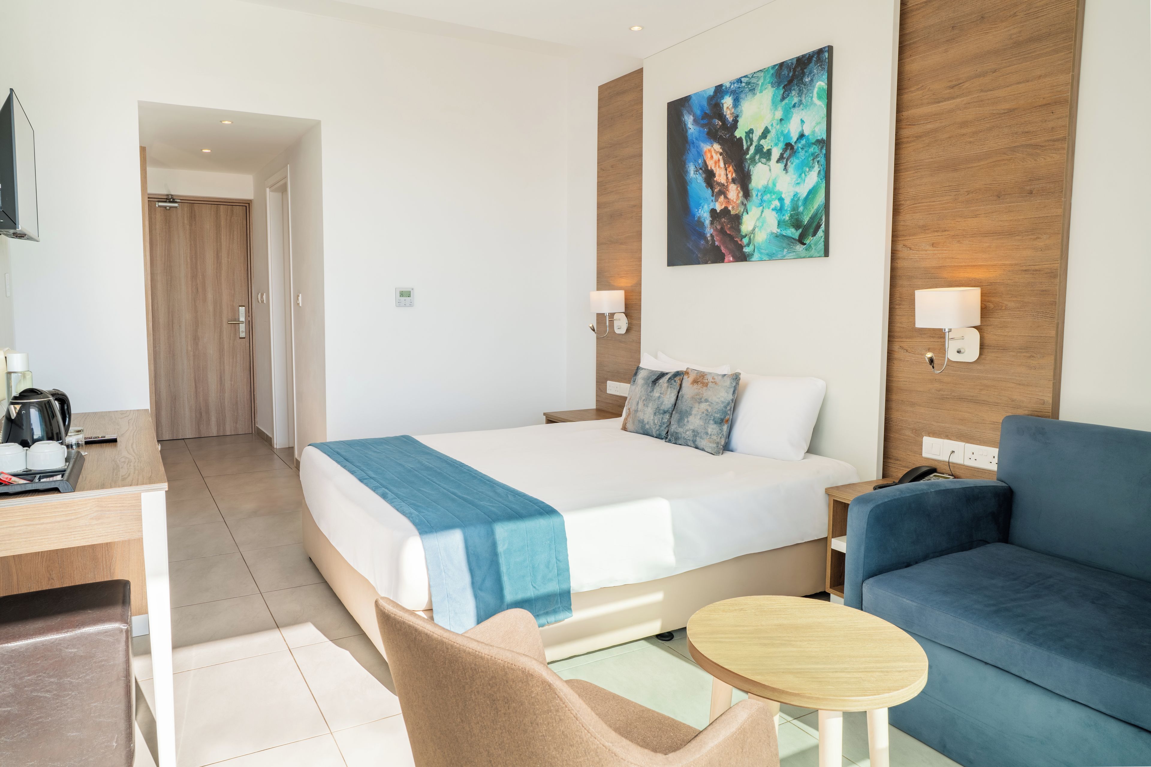 Una habitación de hotel con una cama, un sofá azul, una mesita redonda y un cuadro en la pared.