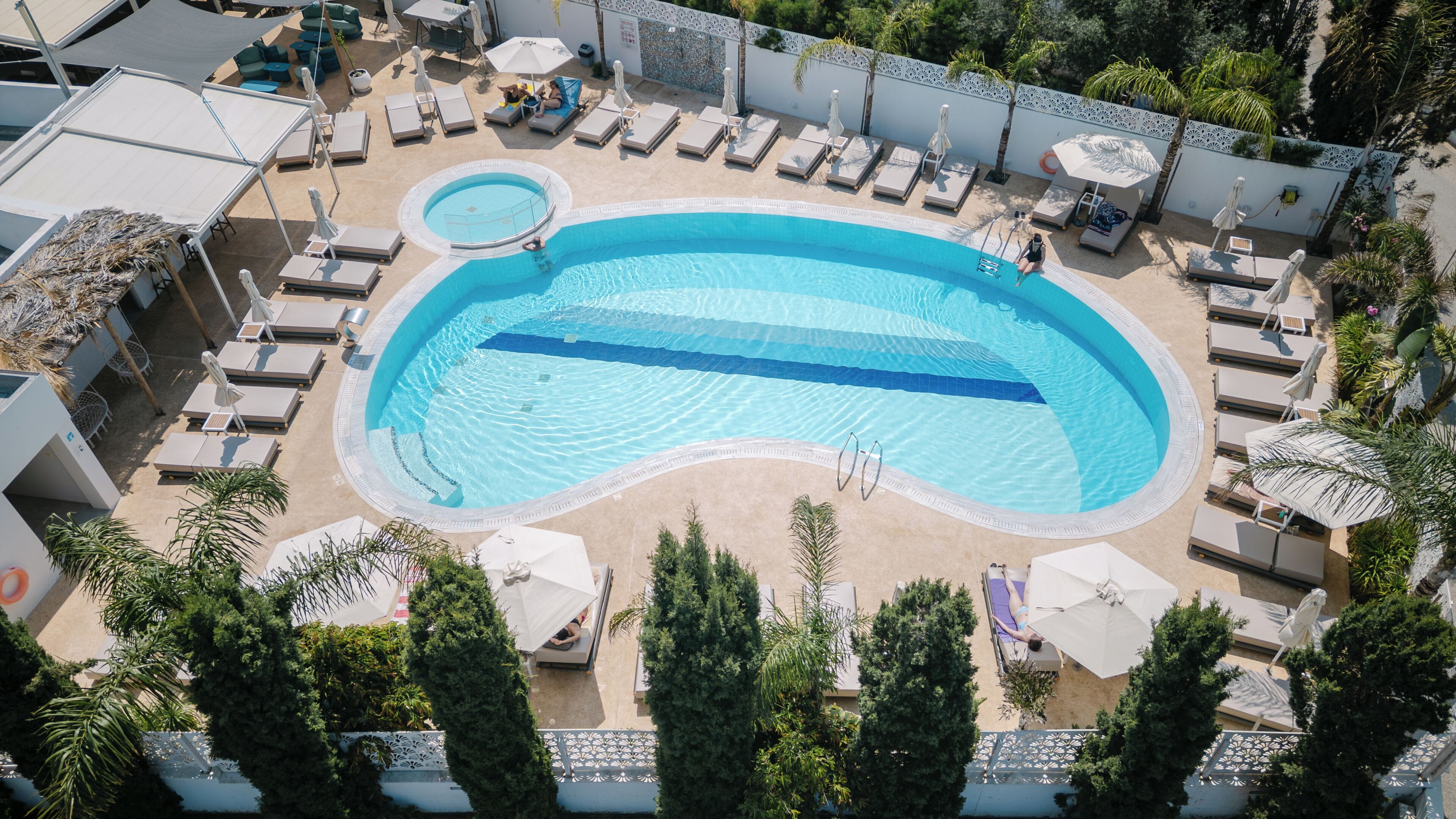 Una piscina en la azotea con varios sillones de sol y sombrillas, rodeada de árboles y una cerca decorativa.