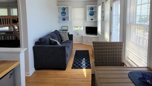 A modern living room with a grey sofa, a small table, and a TV. There are decorative shelves with blue items and a ceiling fan. - Water View " THE PIPERS NEST " - minutes to the beach, located in Snug harbour ! (South Kingstown)