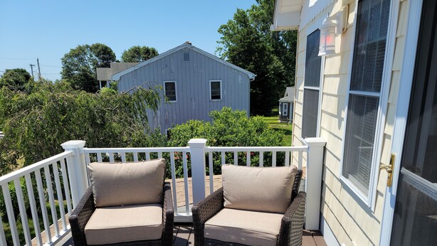 A covered outdoor seating area with wicker chairs and a small table, a white railing, and a view of a house in the background. - Water View " THE PIPERS NEST " - minutes to the beach, located in Snug harbour ! (South Kingstown)