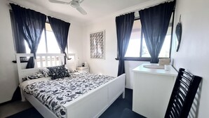 Ein Schlafzimmer mit einem Bett, einem Schreibtisch, einem Stuhl, einem Deckenventilator und einem Fenster mit Vorhängen.