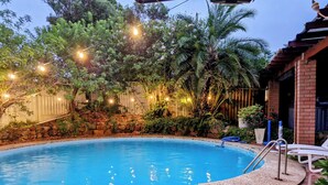 Ein Swimmingpool, umgeben von einem Garten mit Lichterketten, einer Steinmauer und einer Pflasterterrasse.