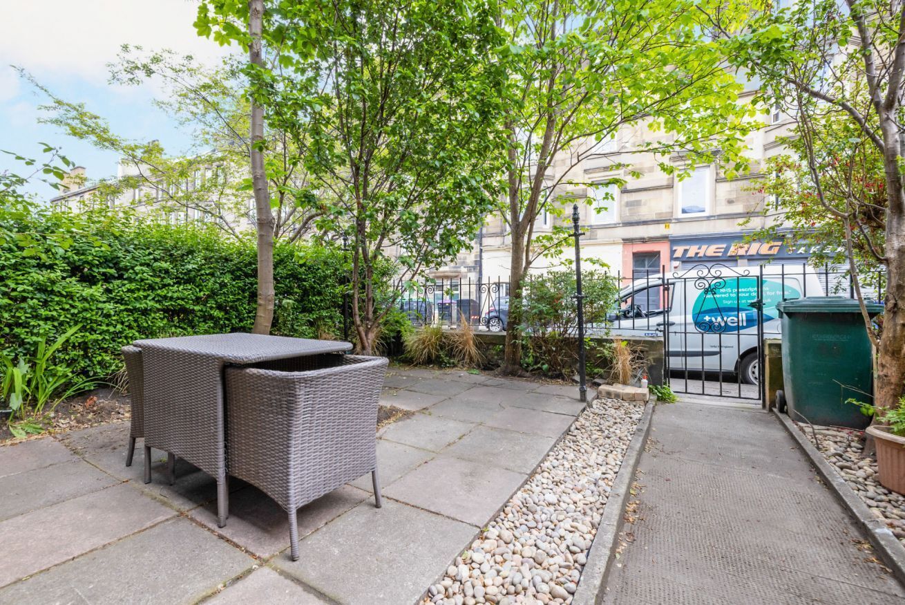 Un espace extérieur aménagé avec une table et des chaises en osier, entouré de verdure et bordé d’un chemin menant à un bâtiment sur lequel est apposée une enseigne indiquant « THE RIG ».