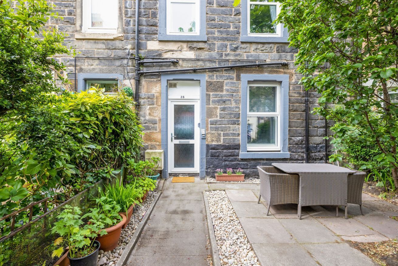 A stone building with a door marked '28', a small garden with potted plants, and a wicker table with chairs.
