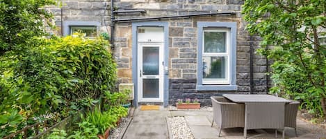 A stone building with a door marked '28', a small garden with potted plants, and a wicker table with chairs.