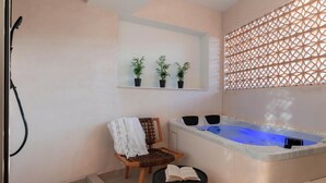 Ein Spa-Raum mit Whirlpool, einem Sessel mit Handtuch, einem kleinen Tisch mit einem Buch und drei Blumentöpfen auf einem Regal.