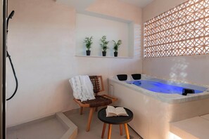 A spa room with a hot tub, a chair with a towel, a small table with a book, and three potted plants on a shelf.