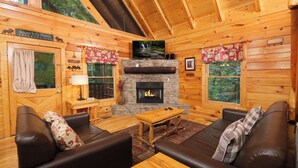 Un intérieur de chalet chaleureux avec une cheminée, des murs en bois, des canapés en cuir et un téléviseur.
