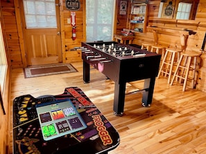 Um quarto com piso de madeira, uma mesa de pebolim, capa para mesa de pebolim e um extintor de incêndio.