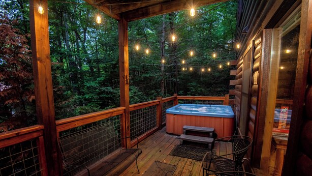 Une terrasse en bois avec un jacuzzi, des guirlandes lumineuses et des chaises donnant sur une forêt.