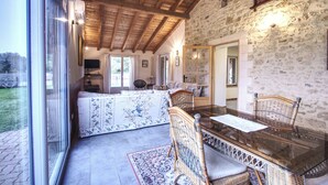 A spacious living area with a glass dining table, wicker chairs, a sofa set, and a stone wall.