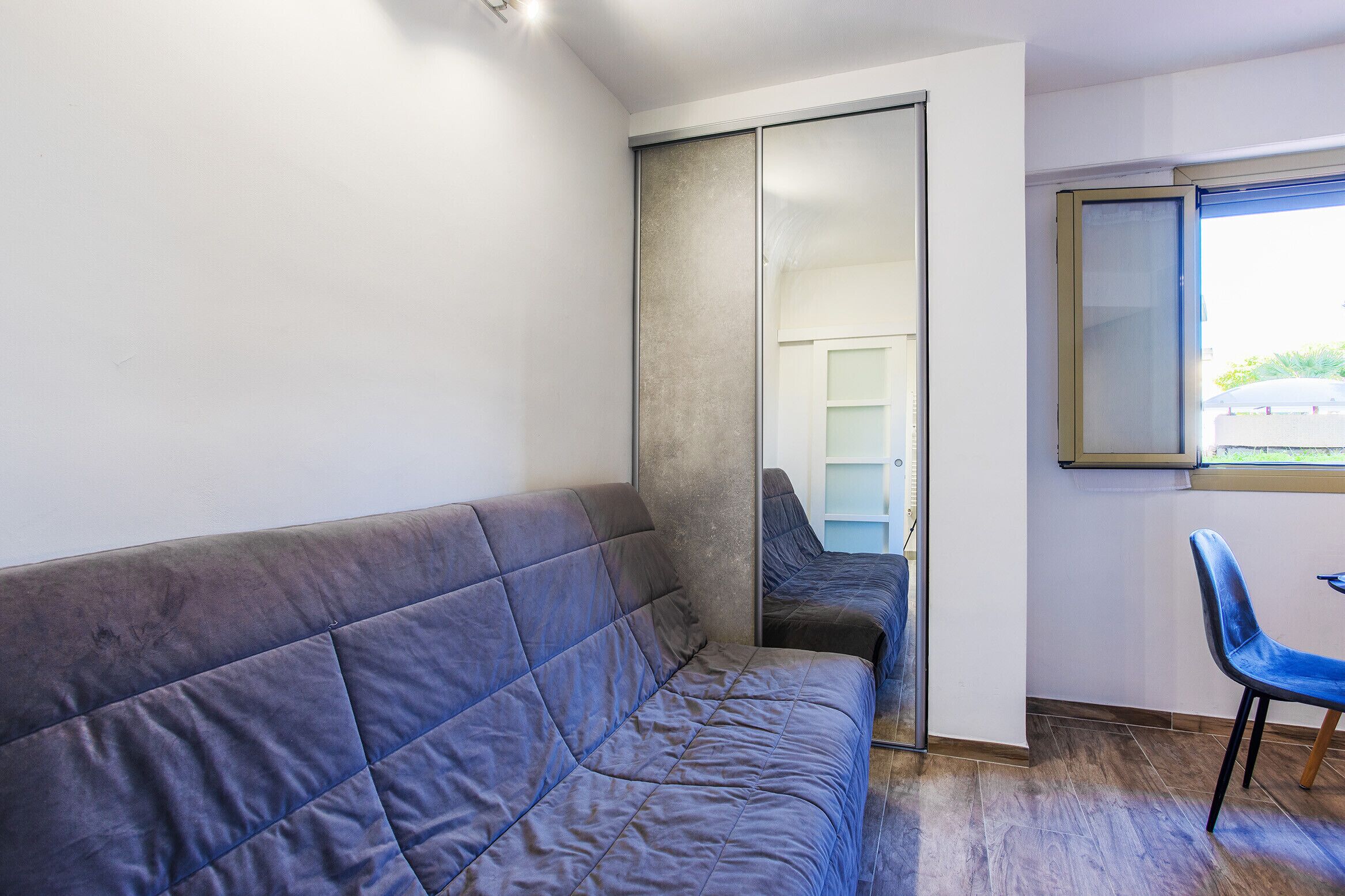 A room with a grey sofa, a blue chair, a sliding door, and a window with a view of greenery outside.