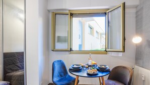 Un petit coin repas avec une table ronde, deux chaises bleues et une fenêtre dont les volets sont ouverts.