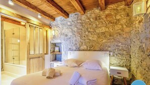A bedroom with a stone wall, wooden ceiling beams, a bed with pillows, a nightstand, and a wall-mounted lamp.
