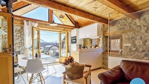 A cozy interior with a stone wall, wooden ceiling, a dining area with a table and chairs, a kitchen with white cabinets, and a view through large windows.