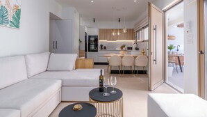 Una sala moderna con un sofá, un comedor con sillas, una cocina al fondo y una puerta que da a otra habitación.