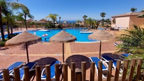 Un espace piscine doté de nombreuses chaises longues et de parasols en chaume, entouré de palmiers et offrant une vue dégagée sur l’océan.