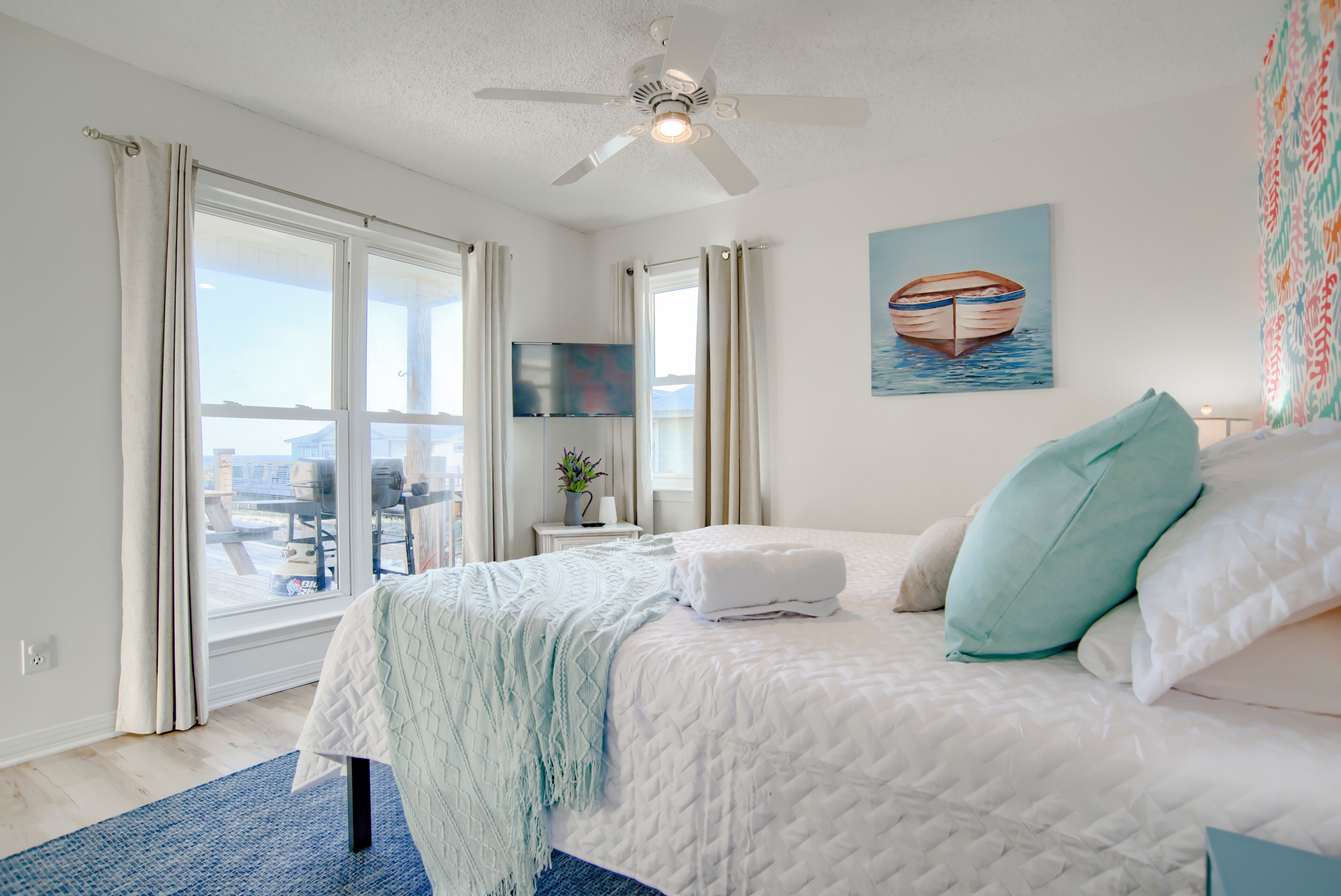 A bedroom with a bed, a ceiling fan, a window with curtains, a painting of a boat, and a view of the outside.