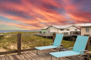 A beachside deck with two lounge chairs, a wooden railing, and a view of beach houses under a colorful sunset sky. - Gulf View bungalow with Hot Tub and Mini Golf (Gulf Shores)