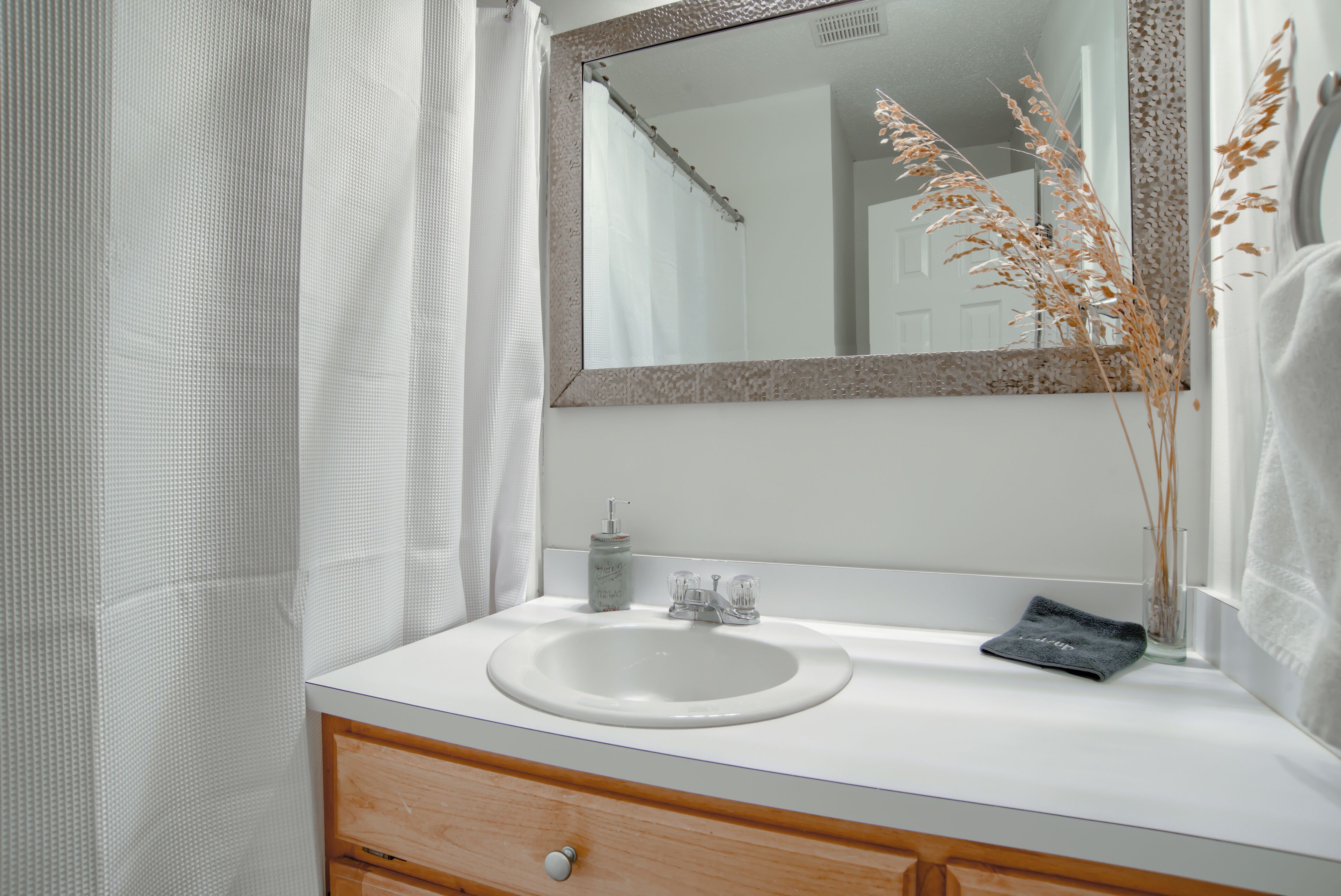 A bathroom with a white sink, a mirror, a wooden cabinet, and a glass vase with dried plants.