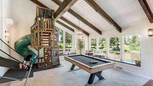 Un vaste espace intérieur comprenant une table de billard, une structure de jeu en bois et de grandes fenêtres donnant sur l’extérieur.
