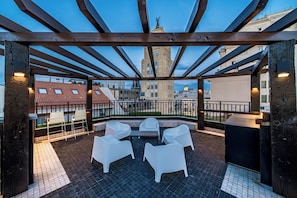 A rooftop terrace with a wooden pergola, white plastic chairs, and a view of city buildings.