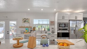 A modern kitchen with white cabinetry, a built-in oven, microwave, and refrigerator. There is a window with a view of the outside, and a countertop with a bowl of fruit and a cutting board. - Catalina Hideaway - Heated Pool - Game Room (Indian Harbour Beach)
