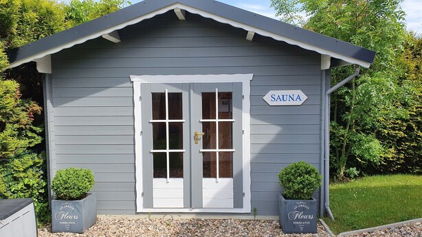 A small building with a sign reading "SAUNA" and two potted plants labeled "LE JARDIN FLEURS SOIRÉE D'ÉTÉ PARIS 1900".