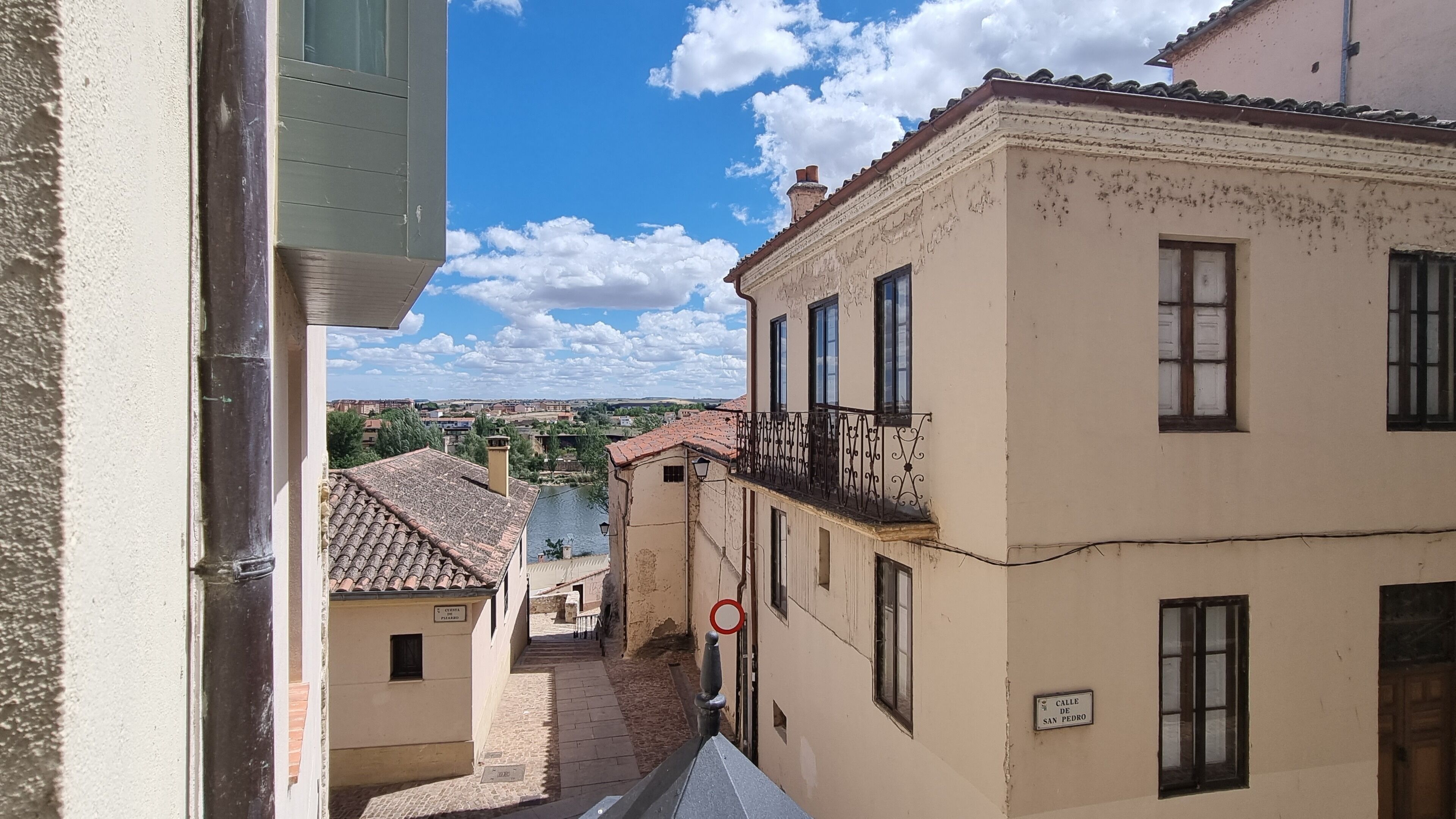 A narrow street with buildings, a clear sky, and a river in the distance.