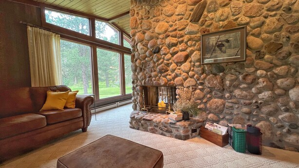 A cozy living room with a stone fireplace, a leather sofa, and a large window offering a view of the outdoors. - Secluded Setting On French Creek In The Southern Black Hills (Custer)