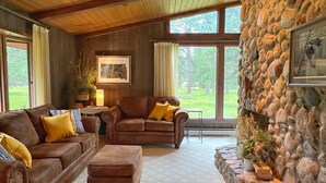 A cozy living room with a stone fireplace, leather furniture, and large windows offering a view of the outdoors. - Secluded Setting On French Creek In The Southern Black Hills (Custer)