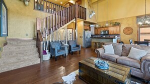 Un vaste espace de vie comprenant un escalier, des canapés en cuir, une table basse en bois et une cuisine à l’arrière-plan.