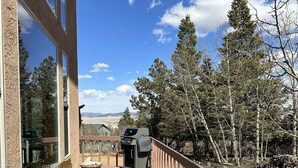 Une terrasse en bois avec un banc et un barbecue, offrant une vue sur une zone boisée et sur des montagnes lointaines.
